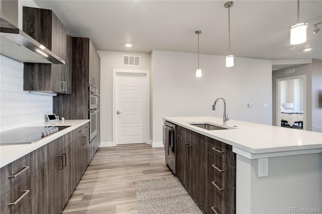 kitchen featuring a center island with sink, black electric cooktop, hanging light fixtures, sink, and wall chimney range hood