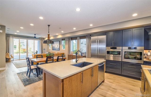 kitchen featuring stainless steel appliances, sink, decorative light fixtures, light hardwood / wood-style flooring, and an island with sink