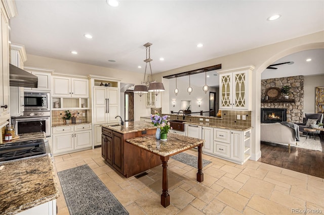 kitchen with ventilation hood, appliances with stainless steel finishes, tasteful backsplash, light stone counters, and a fireplace