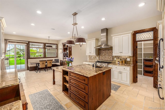 kitchen featuring wall chimney exhaust hood, backsplash, hanging light fixtures, a kitchen island with sink, and light stone countertops