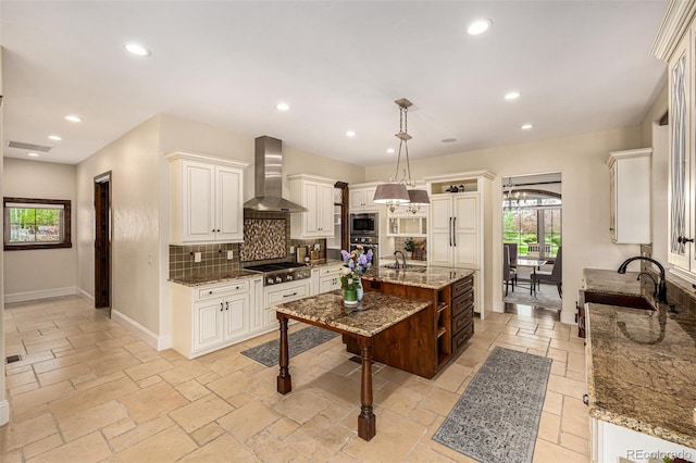 kitchen with light stone countertops, hanging light fixtures, backsplash, appliances with stainless steel finishes, and wall chimney range hood