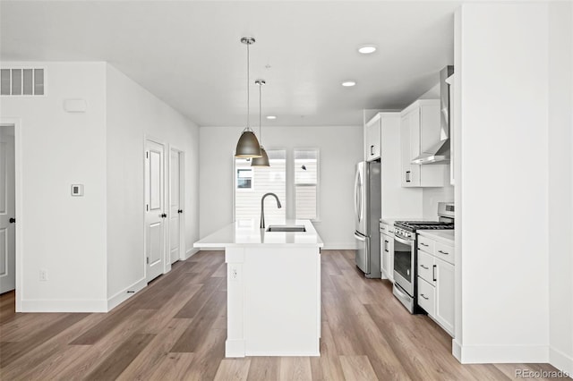 kitchen with sink, hanging light fixtures, an island with sink, stainless steel appliances, and wall chimney range hood