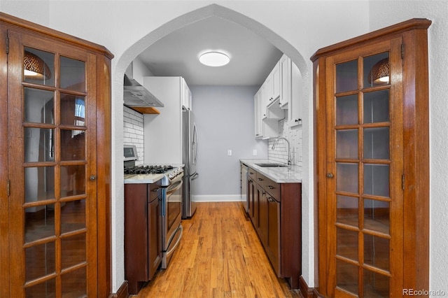 kitchen with stainless steel appliances, a sink, wall chimney range hood, light wood-type flooring, and decorative backsplash