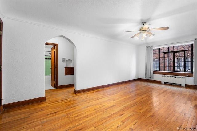 empty room with arched walkways, radiator, light wood-style flooring, a ceiling fan, and baseboards