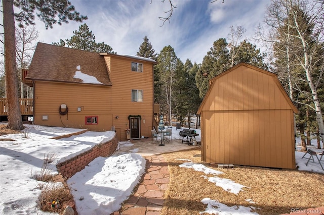 view of snow covered exterior featuring a shingled roof, an outbuilding, a patio, and a storage unit