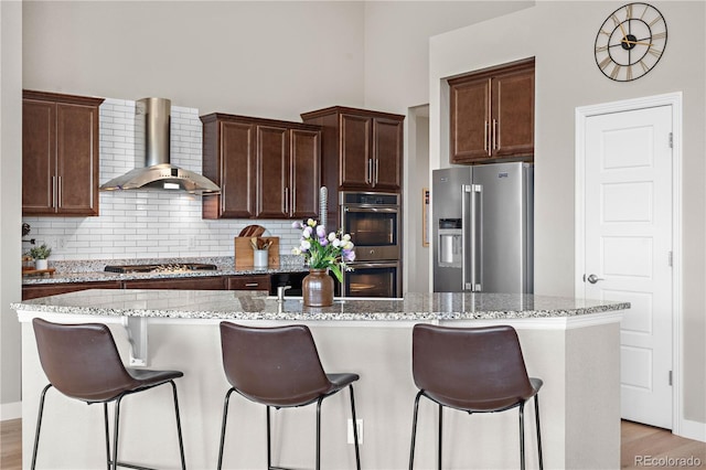 kitchen featuring a kitchen island with sink, backsplash, appliances with stainless steel finishes, light stone counters, and wall chimney exhaust hood