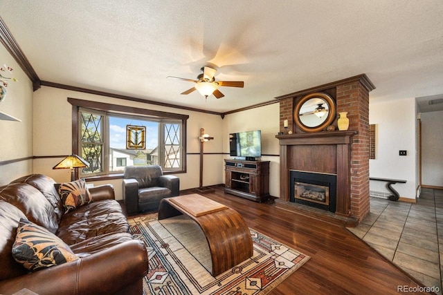 living area with a textured ceiling, crown molding, a brick fireplace, ceiling fan, and wood finished floors