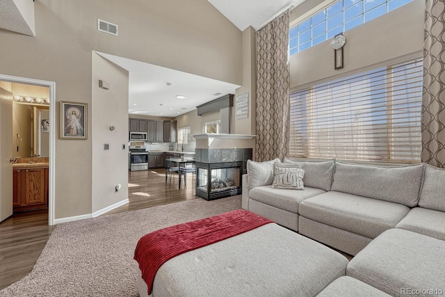 living area featuring high vaulted ceiling, wood finished floors, a multi sided fireplace, visible vents, and baseboards