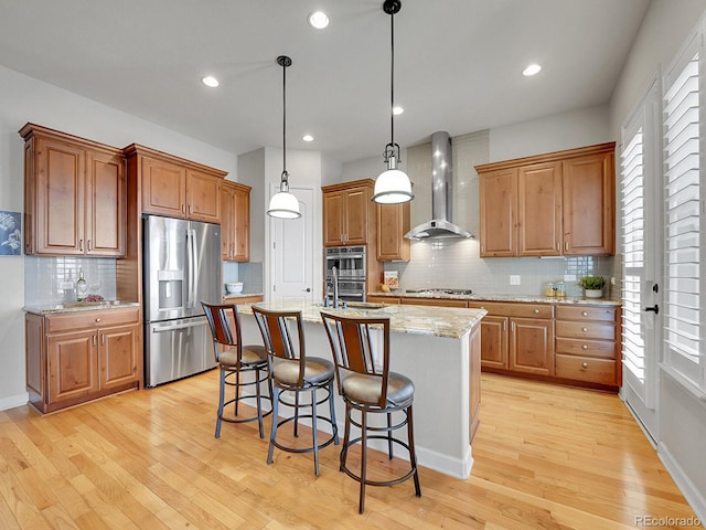 kitchen featuring a kitchen island with sink, stainless steel appliances, light stone countertops, decorative light fixtures, and wall chimney exhaust hood