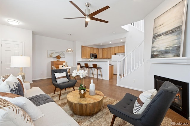 living area featuring baseboards, a glass covered fireplace, wood finished floors, stairway, and ceiling fan