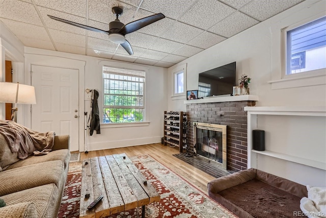 living room with ceiling fan, a drop ceiling, a brick fireplace, and light hardwood / wood-style flooring