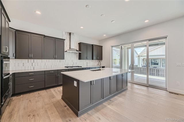 kitchen with light hardwood / wood-style flooring, wall chimney exhaust hood, sink, an island with sink, and stainless steel gas cooktop