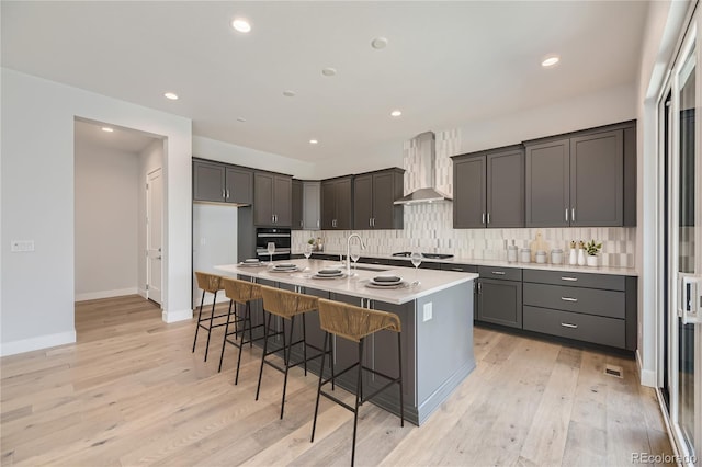 kitchen featuring sink, wall chimney range hood, a center island with sink, light hardwood / wood-style floors, and black oven