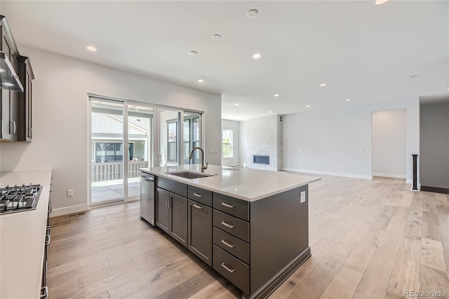 kitchen with appliances with stainless steel finishes, a stone fireplace, sink, a kitchen island with sink, and light hardwood / wood-style flooring