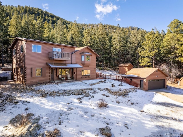 snow covered rear of property featuring a forest view, a detached garage, a balcony, and an outdoor structure