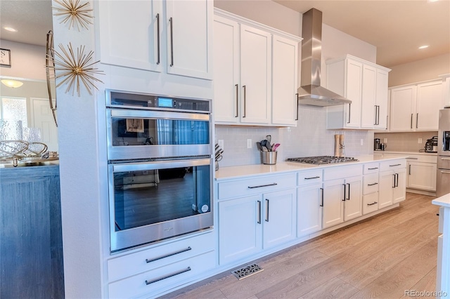 kitchen featuring stainless steel appliances, wall chimney range hood, tasteful backsplash, light hardwood / wood-style flooring, and white cabinets