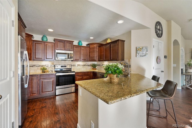 kitchen with light stone countertops, stainless steel appliances, a kitchen breakfast bar, dark hardwood / wood-style flooring, and backsplash