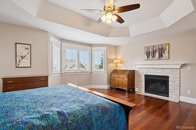 bedroom with ceiling fan, a fireplace, wood finished floors, baseboards, and a tray ceiling