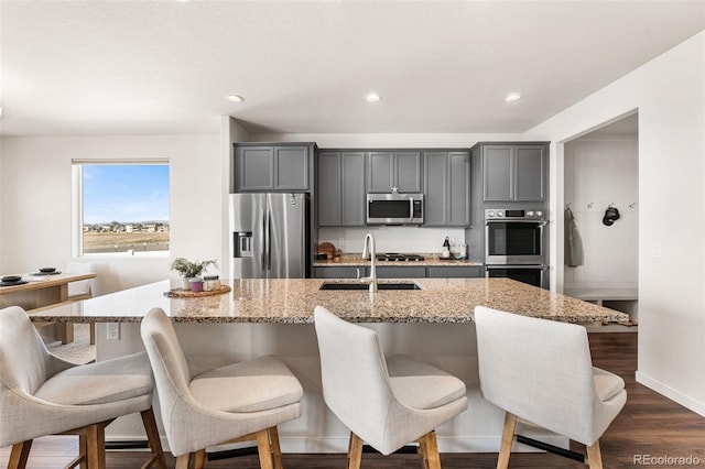 kitchen featuring a kitchen bar, a kitchen island with sink, gray cabinetry, appliances with stainless steel finishes, and dark wood-style floors