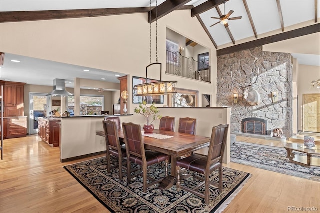 dining room featuring beam ceiling, ceiling fan, a stone fireplace, and light wood-type flooring