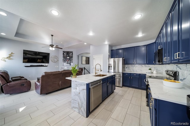 kitchen with blue cabinetry, stainless steel appliances, a tray ceiling, light stone countertops, and a center island with sink