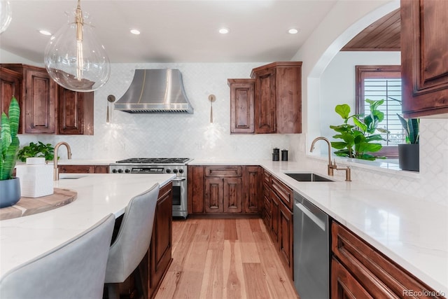 kitchen with stainless steel appliances, sink, wall chimney range hood, light hardwood / wood-style floors, and hanging light fixtures