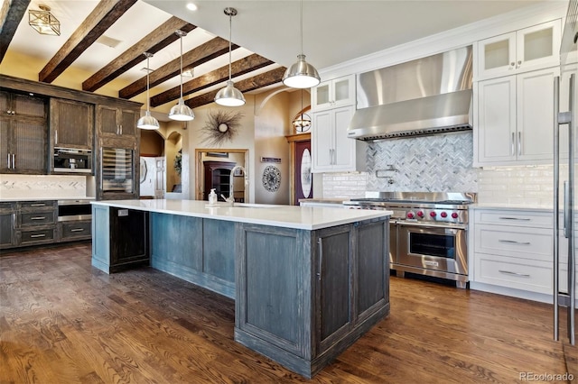 kitchen featuring appliances with stainless steel finishes, decorative backsplash, a kitchen island with sink, wall chimney range hood, and beam ceiling