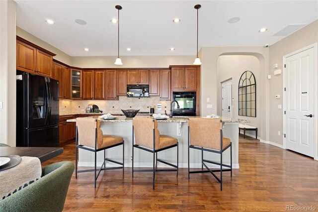 kitchen with light stone counters, decorative light fixtures, black appliances, an island with sink, and glass insert cabinets