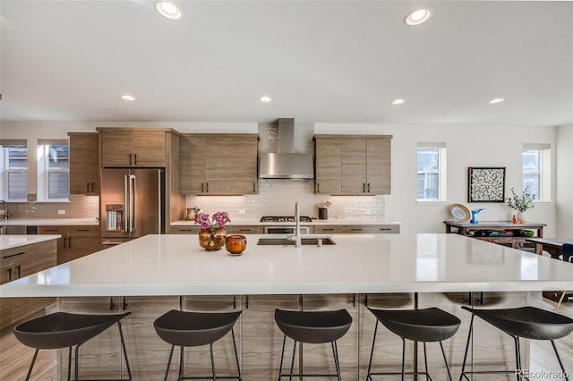 kitchen with a breakfast bar area, high end refrigerator, a sink, wall chimney range hood, and brown cabinetry