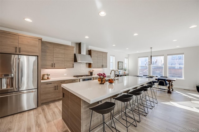 kitchen featuring backsplash, appliances with stainless steel finishes, light wood-style floors, wall chimney range hood, and a kitchen breakfast bar