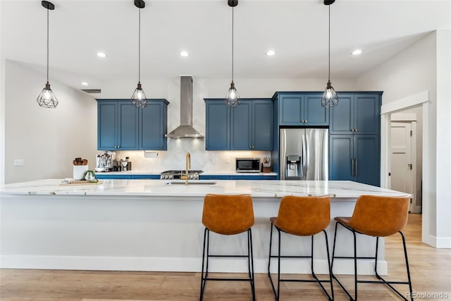kitchen with wall chimney range hood, appliances with stainless steel finishes, a breakfast bar, blue cabinetry, and a spacious island