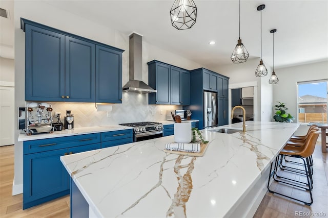 kitchen featuring wall chimney range hood, backsplash, blue cabinetry, appliances with stainless steel finishes, and a sink