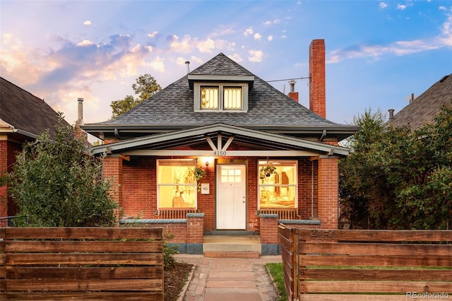 view of front of house featuring a porch, brick siding, fence, and roof with shingles