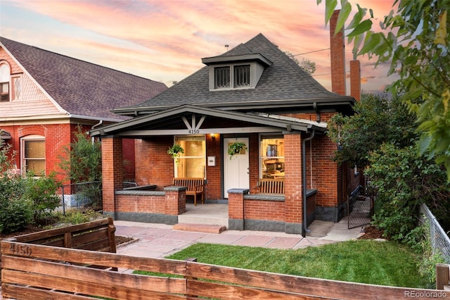 view of front facade featuring covered porch, brick siding, roof with shingles, and fence