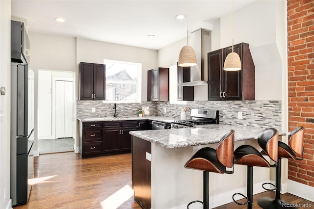 kitchen featuring dark brown cabinetry, a peninsula, stainless steel appliances, light wood-type flooring, and wall chimney range hood