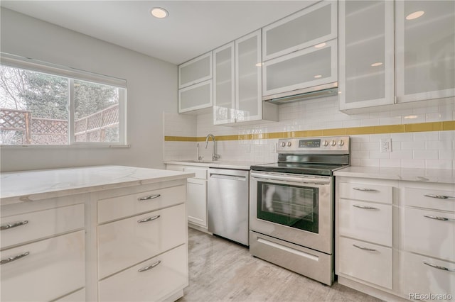 kitchen with light stone counters, stainless steel appliances, a sink, white cabinetry, and glass insert cabinets