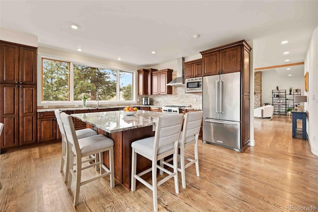 kitchen with appliances with stainless steel finishes, a kitchen island, light wood finished floors, and wall chimney exhaust hood