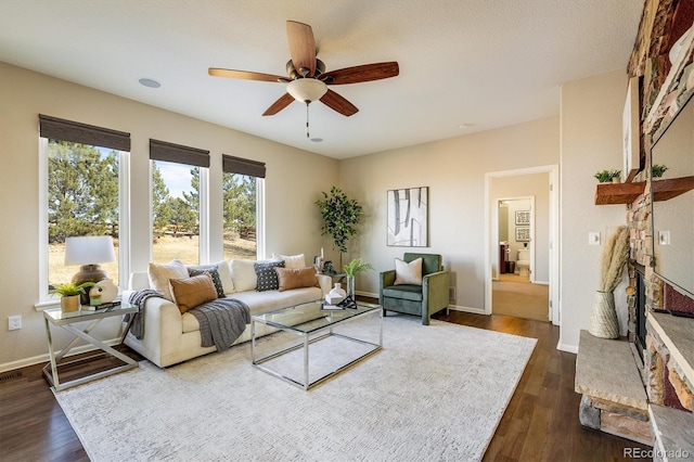 living area with ceiling fan, a stone fireplace, dark wood finished floors, and baseboards