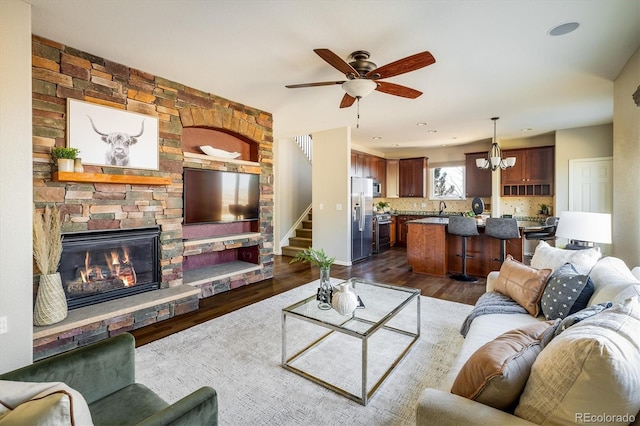 living area featuring stairs, dark wood-style flooring, a fireplace, and ceiling fan with notable chandelier