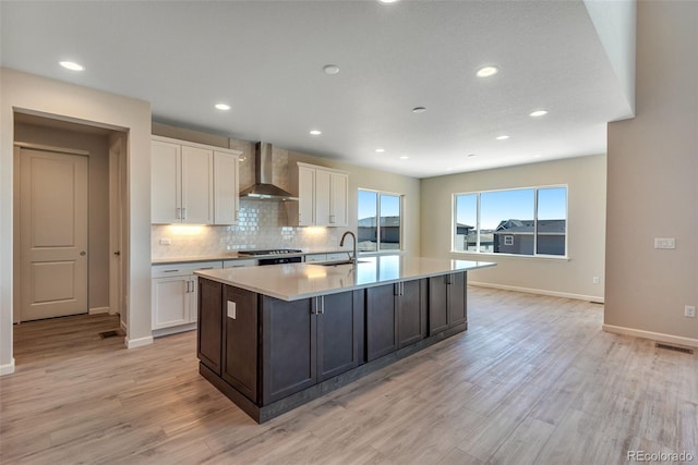 kitchen featuring white cabinets, wall chimney exhaust hood, an island with sink, and light hardwood / wood-style flooring