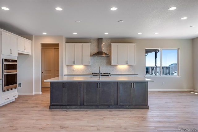 kitchen with an island with sink, light hardwood / wood-style flooring, wall chimney exhaust hood, and white cabinets