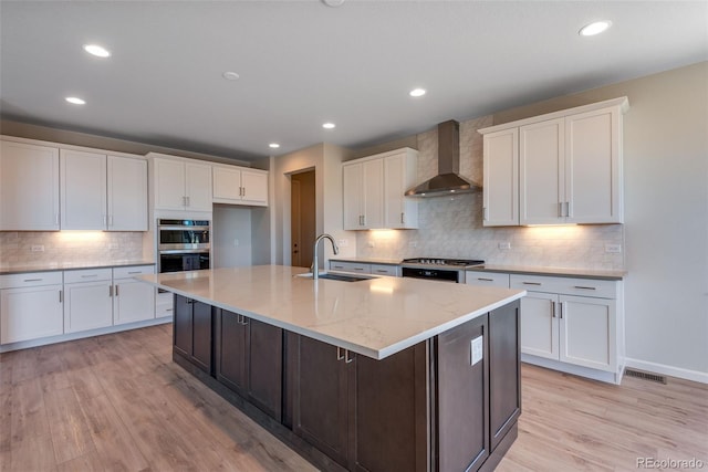 kitchen featuring sink, wall chimney exhaust hood, a kitchen island with sink, light hardwood / wood-style flooring, and white cabinets