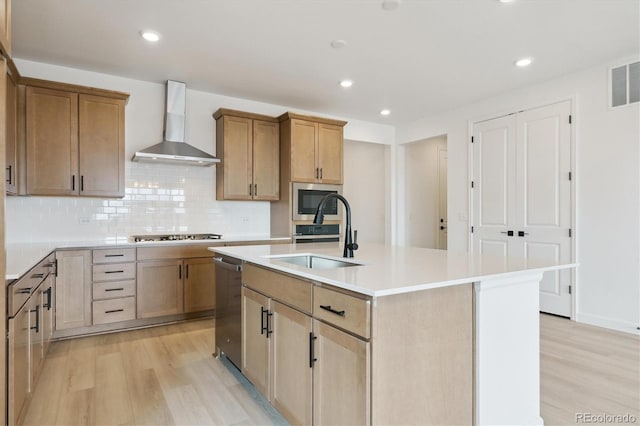 kitchen featuring sink, a kitchen island with sink, stainless steel appliances, wall chimney exhaust hood, and light wood-type flooring