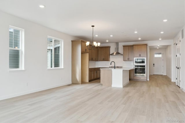 kitchen featuring wall chimney exhaust hood, hanging light fixtures, appliances with stainless steel finishes, a notable chandelier, and a kitchen island with sink