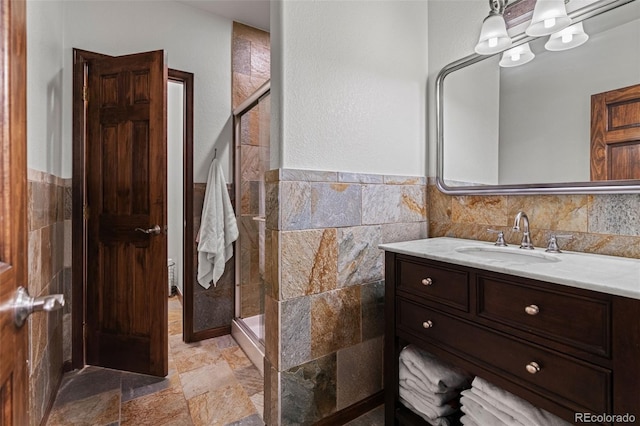 bathroom featuring a stall shower, tile walls, stone finish floors, vanity, and a wainscoted wall