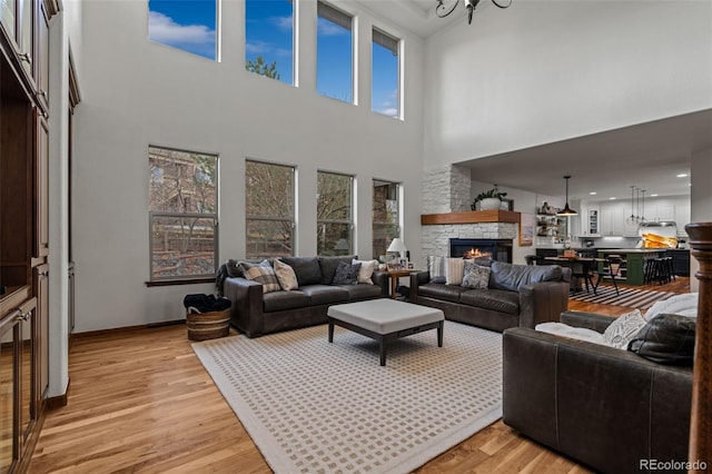 living area featuring a stone fireplace, light wood-type flooring, a towering ceiling, a chandelier, and baseboards