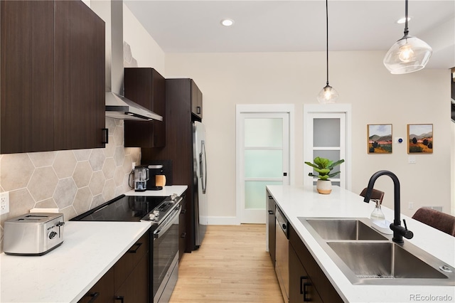 kitchen featuring dark brown cabinets, wall chimney range hood, decorative light fixtures, stainless steel appliances, and a sink