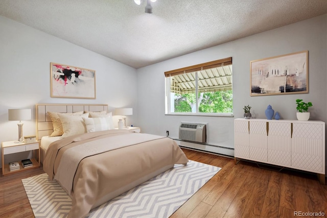 bedroom featuring a textured ceiling, vaulted ceiling, wood finished floors, a baseboard heating unit, and an AC wall unit
