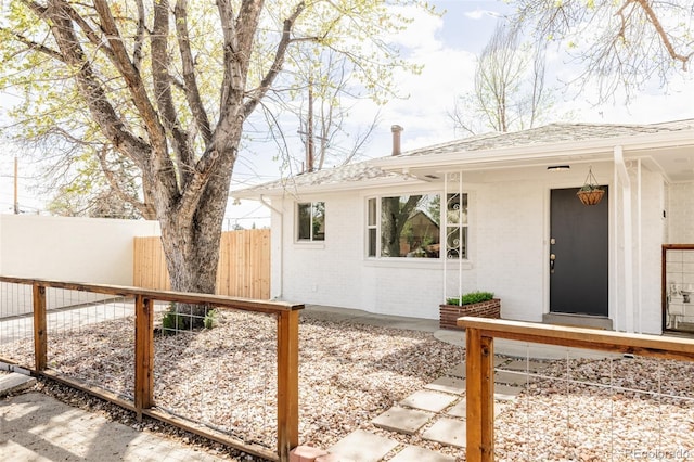 doorway to property featuring fence, a shingled roof, and brick siding