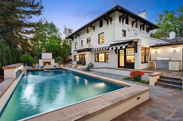 back house at dusk featuring an outdoor fireplace, an outdoor kitchen, a patio area, and pool water feature